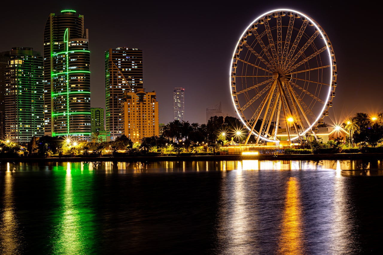 Vibrant night view of Sharjah cityscape featuring a lit Ferris wheel reflecting on water.